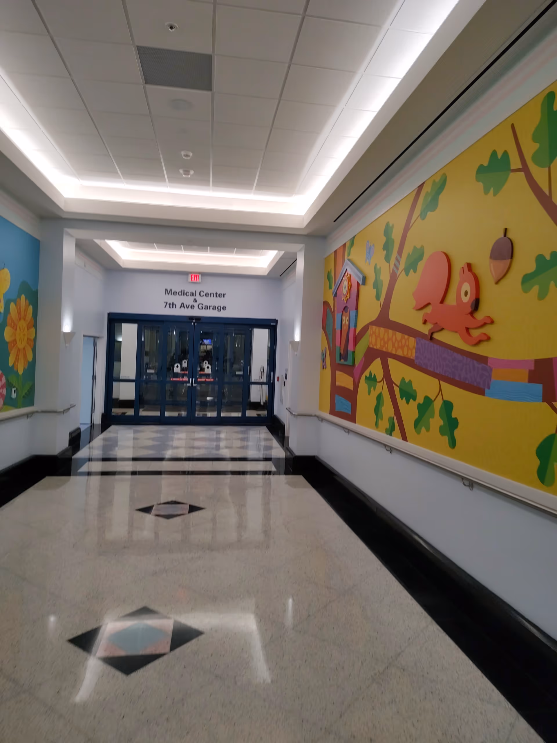 Bright hallway inside a medical facility with a colorful mural and glass doors ahead.