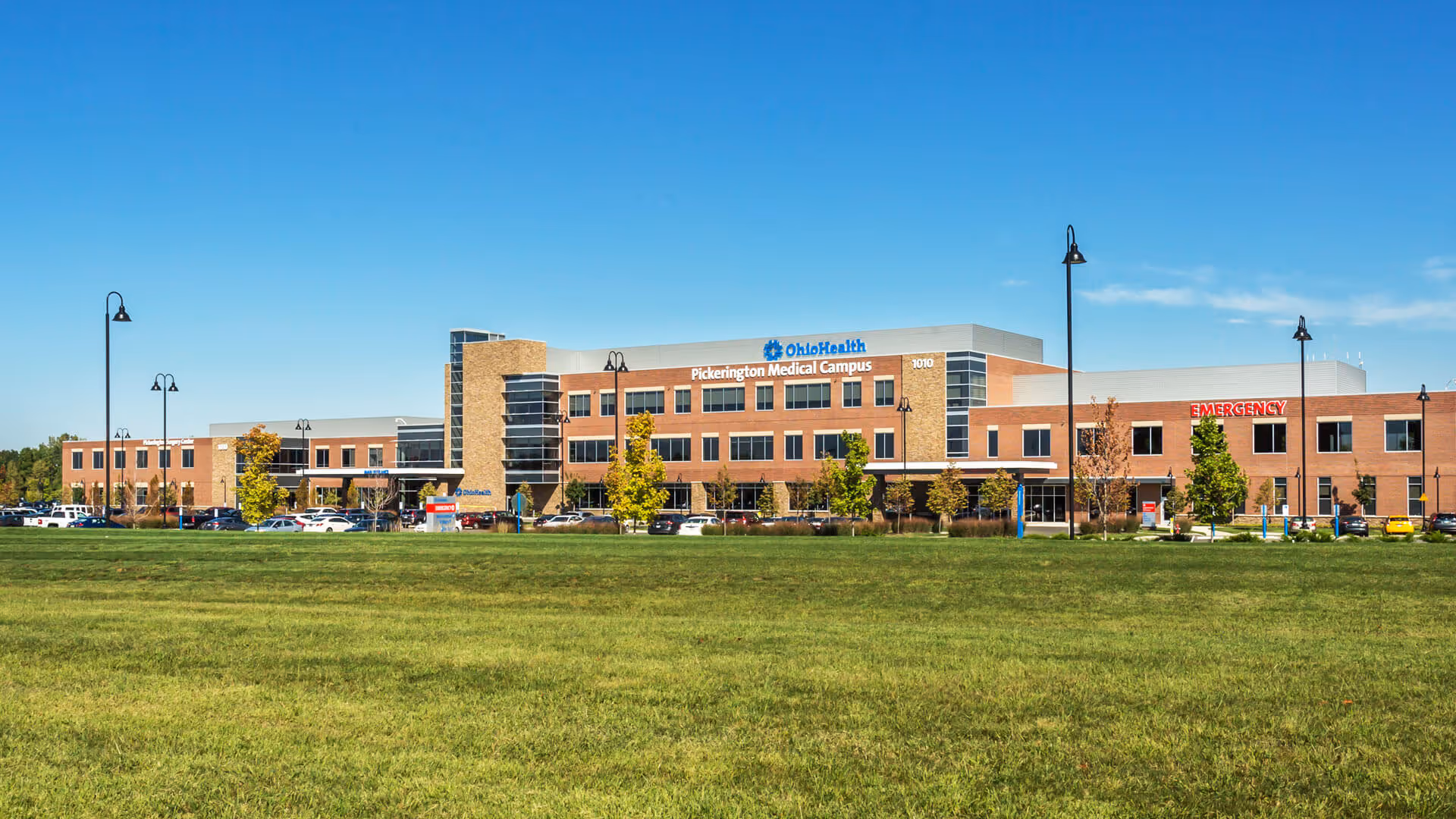 A brick medical campus building with signage, cars parked along a front lot, and grassy foreground.