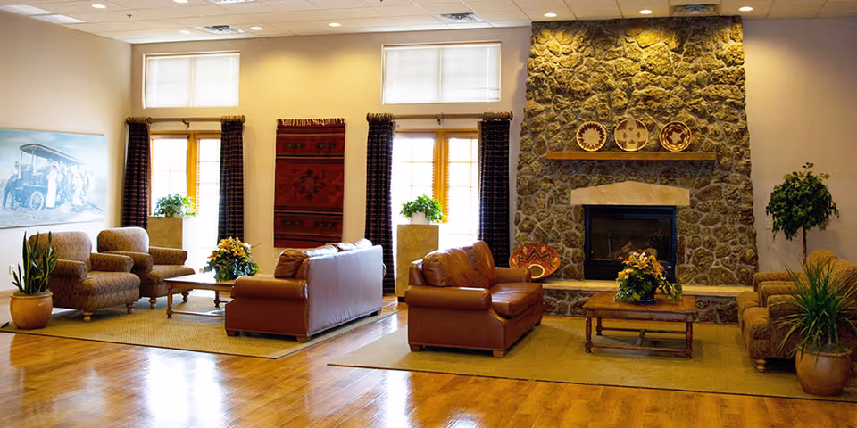 A cozy senior living community lounge with wooden floors, leather sofas, armchairs, a stone fireplace decorated with plates, potted plants, and large windows with curtains allowing natural light.
