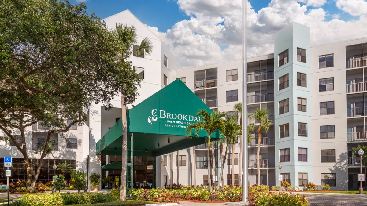 White multi-story senior living building with a green Brookdale entrance canopy and palm trees.