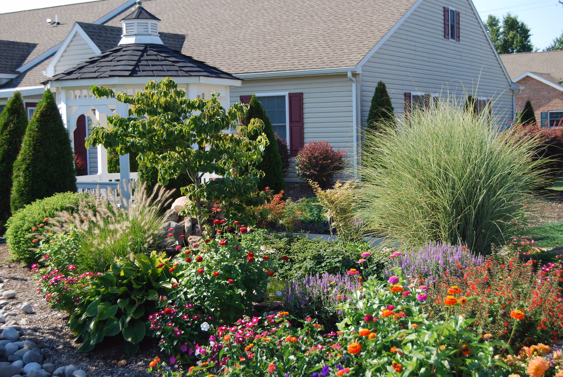 Colorful flower beds and ornamental grasses surrounding a white gazebo in front of a single-story residential building.