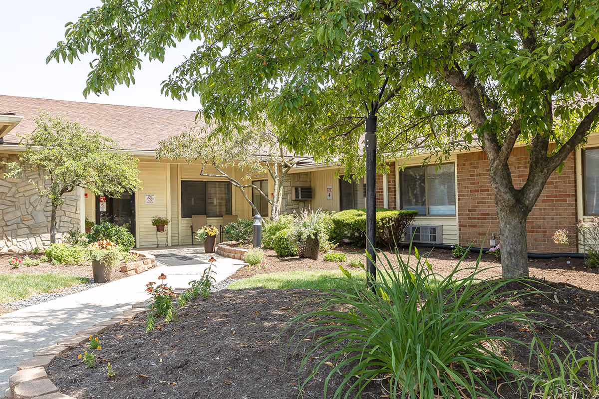 Entrance courtyard and front facade of a senior care building with a walkway, potted plants, trees, and landscaping.