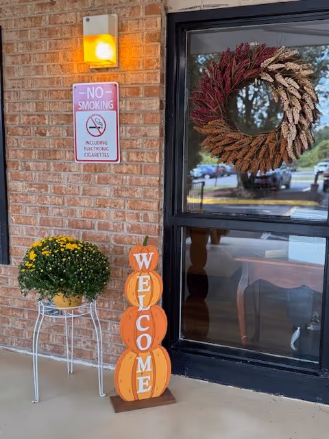 Brick exterior entrance with a wreath, no-smoking sign, pumpkins spelling WELCOME, and potted mums.