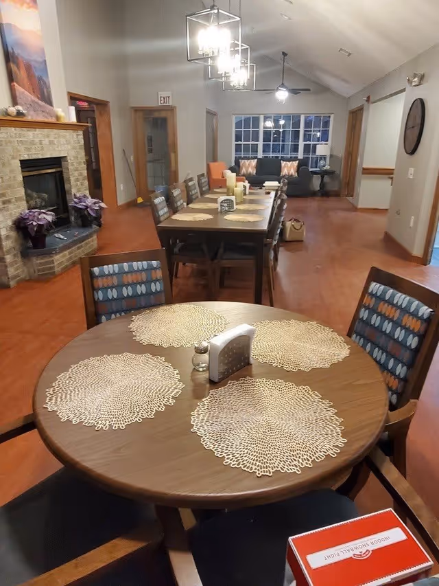 Communal dining room with round and rectangular tables set with placemats, chairs, a fireplace to the left, and a seating area by windows in the background.