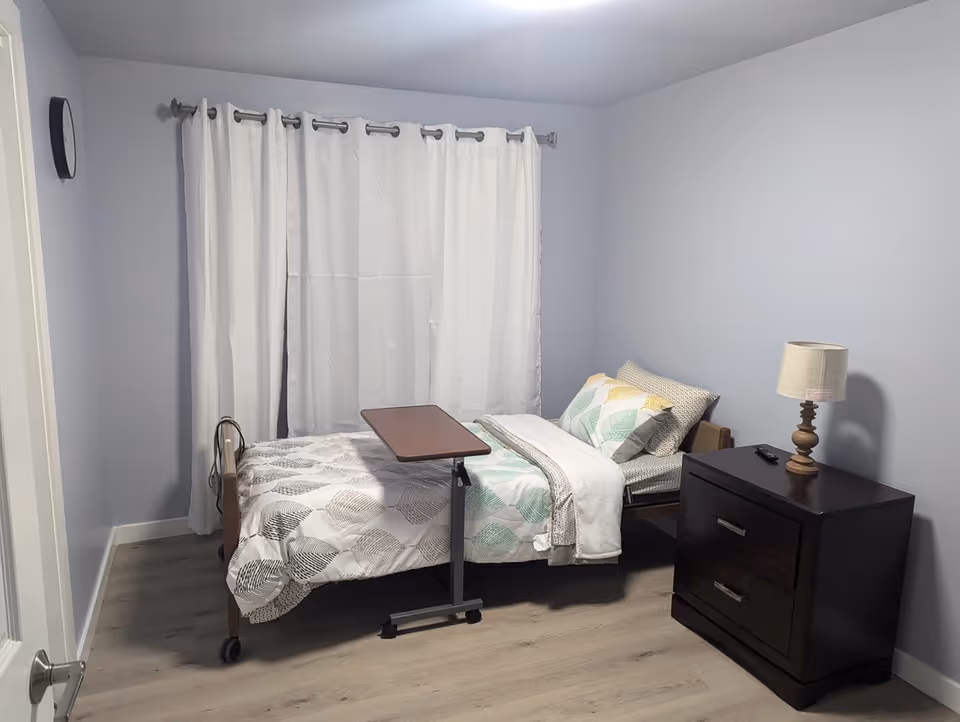 A tidy bedroom with a single bed, nightstand, dresser, lamp, and white curtains.