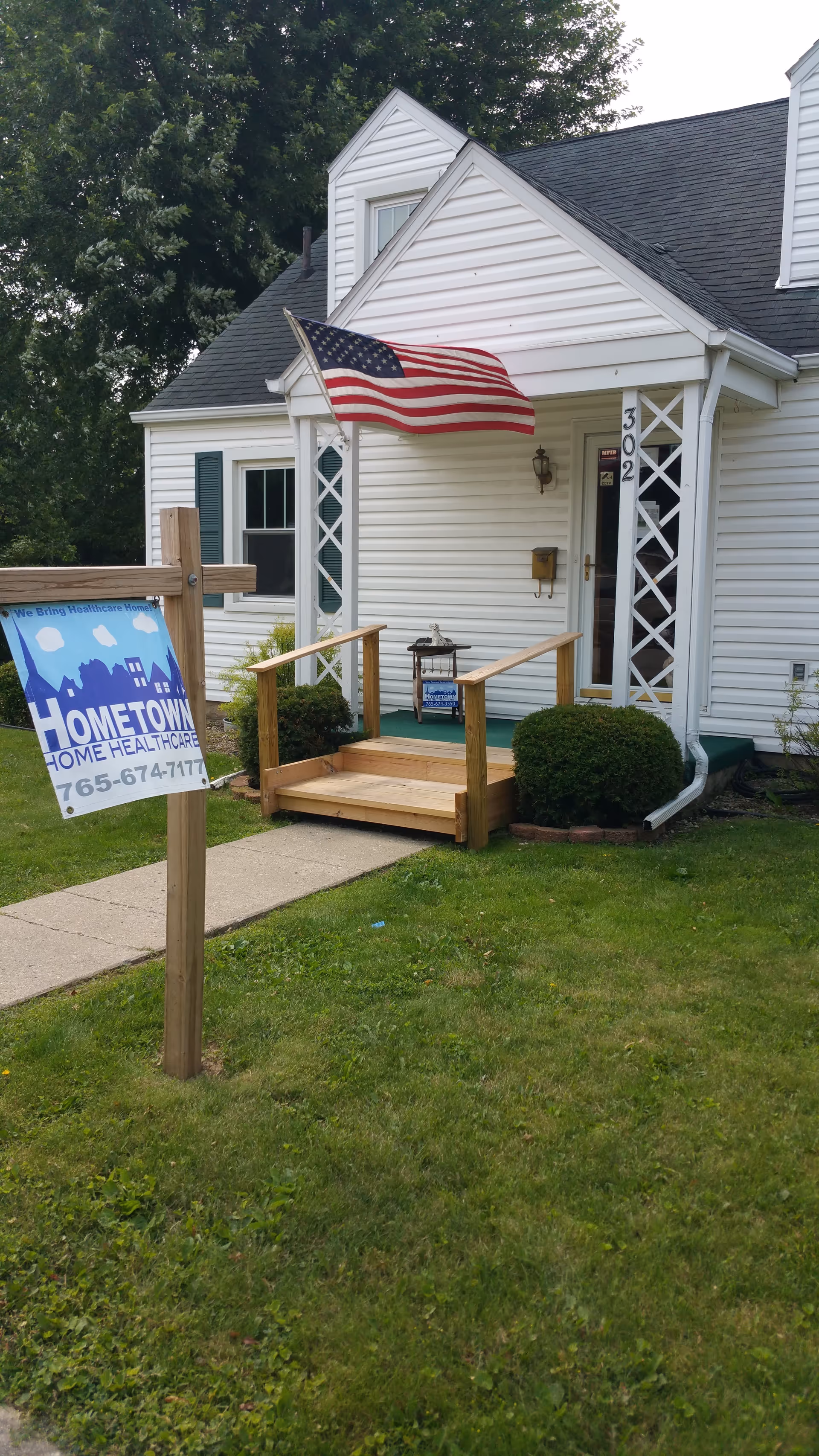 White senior care facility exterior with porch, steps, American flag, and a sign.