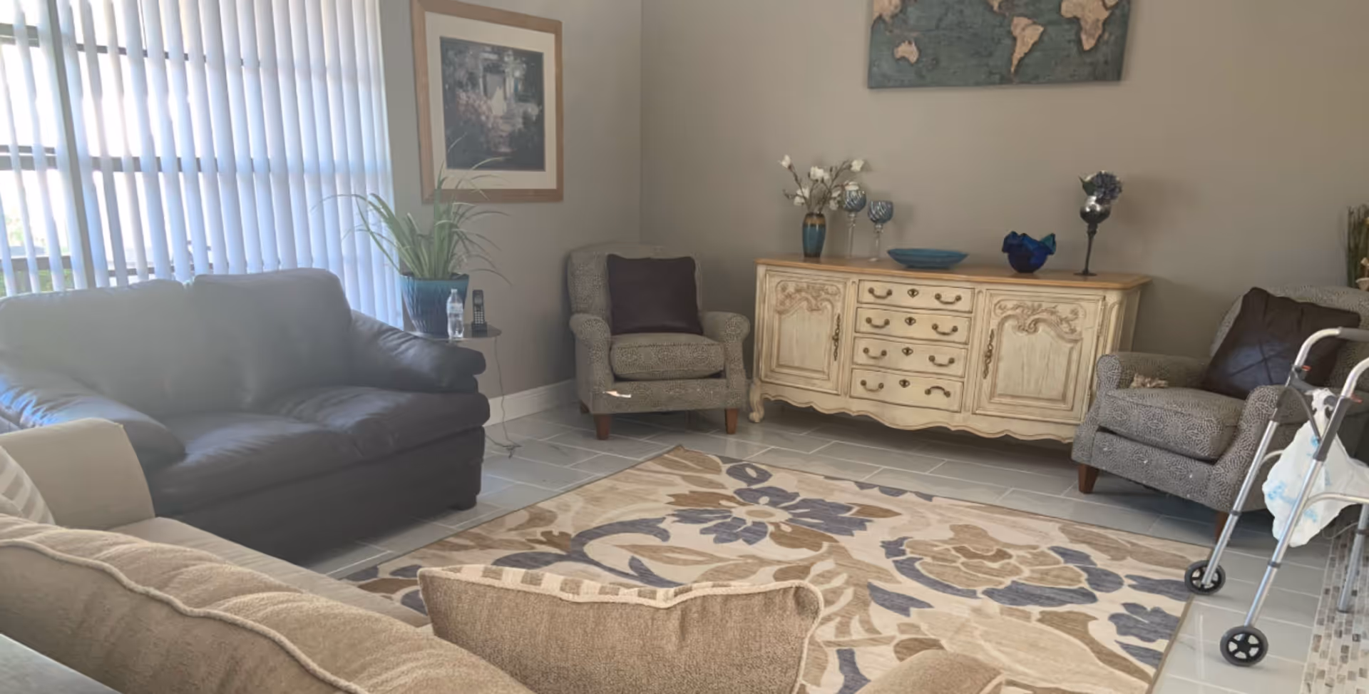 Bright living room with sofas and armchairs around a patterned rug, a decorative sideboard, window blinds, and a walker near a chair.