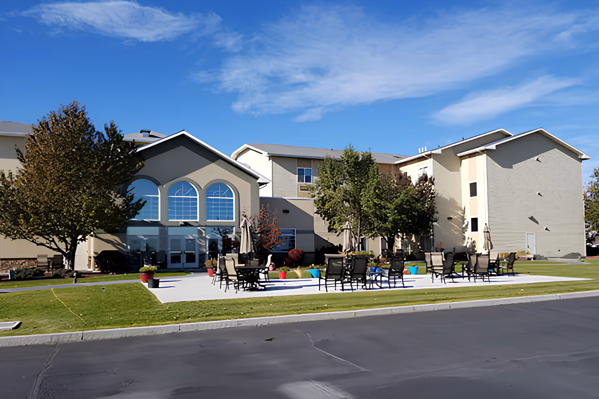 Front exterior of a multi-story senior living facility with lawn and outdoor patio seating under a blue sky.