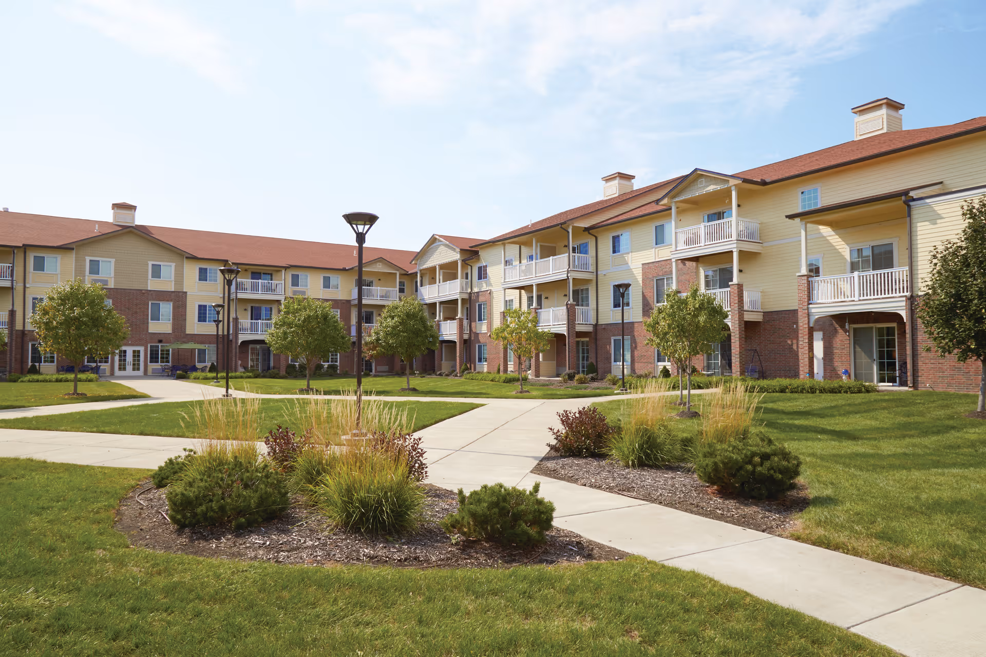 Three-story yellow-and-brick senior living building with balconies surrounding a landscaped central courtyard and walkways.