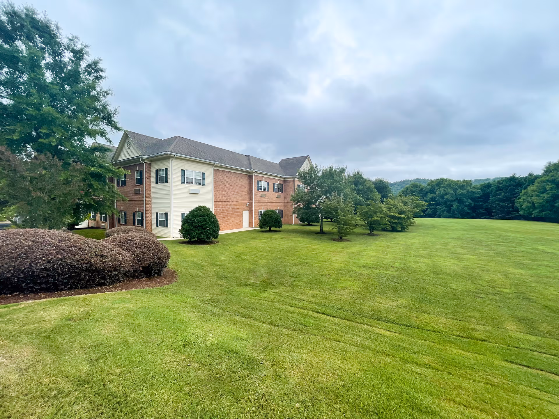 Two-story brick-and-siding senior living building beside a wide green lawn and trees under a cloudy sky.