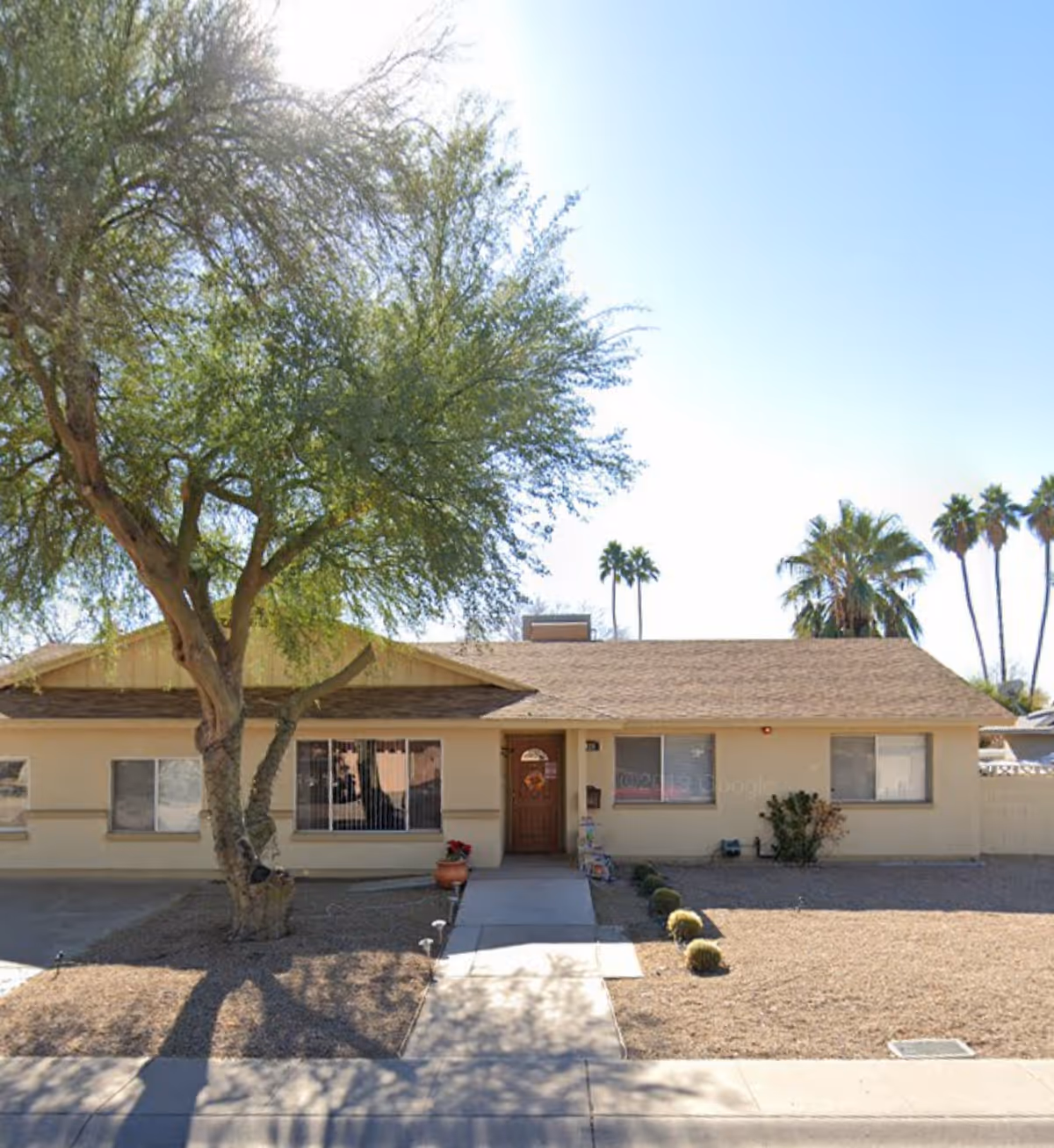 Front exterior of a single-story beige care home with a large tree and desert landscaping.