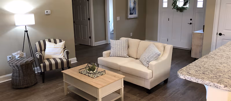 A cozy living room area with a beige loveseat adorned with two patterned pillows, a striped armchair with a white pillow, a wooden coffee table with decorative items, a wicker side table, a floor lamp, and a front door decorated with a green wreath.