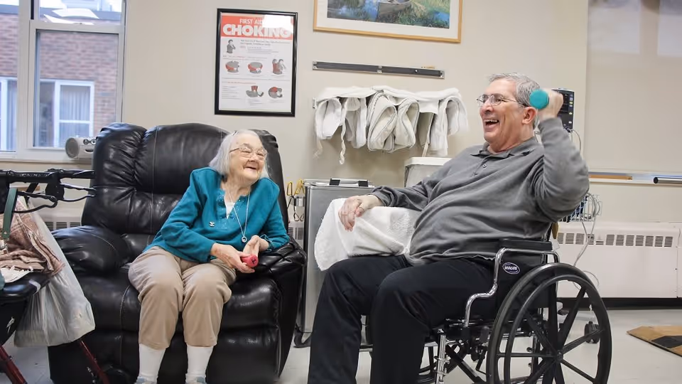 An elderly woman sitting in a black leather recliner and an elderly man in a wheelchair are smiling and exercising with small hand weights in a room with medical equipment and a first aid choking poster on the wall.