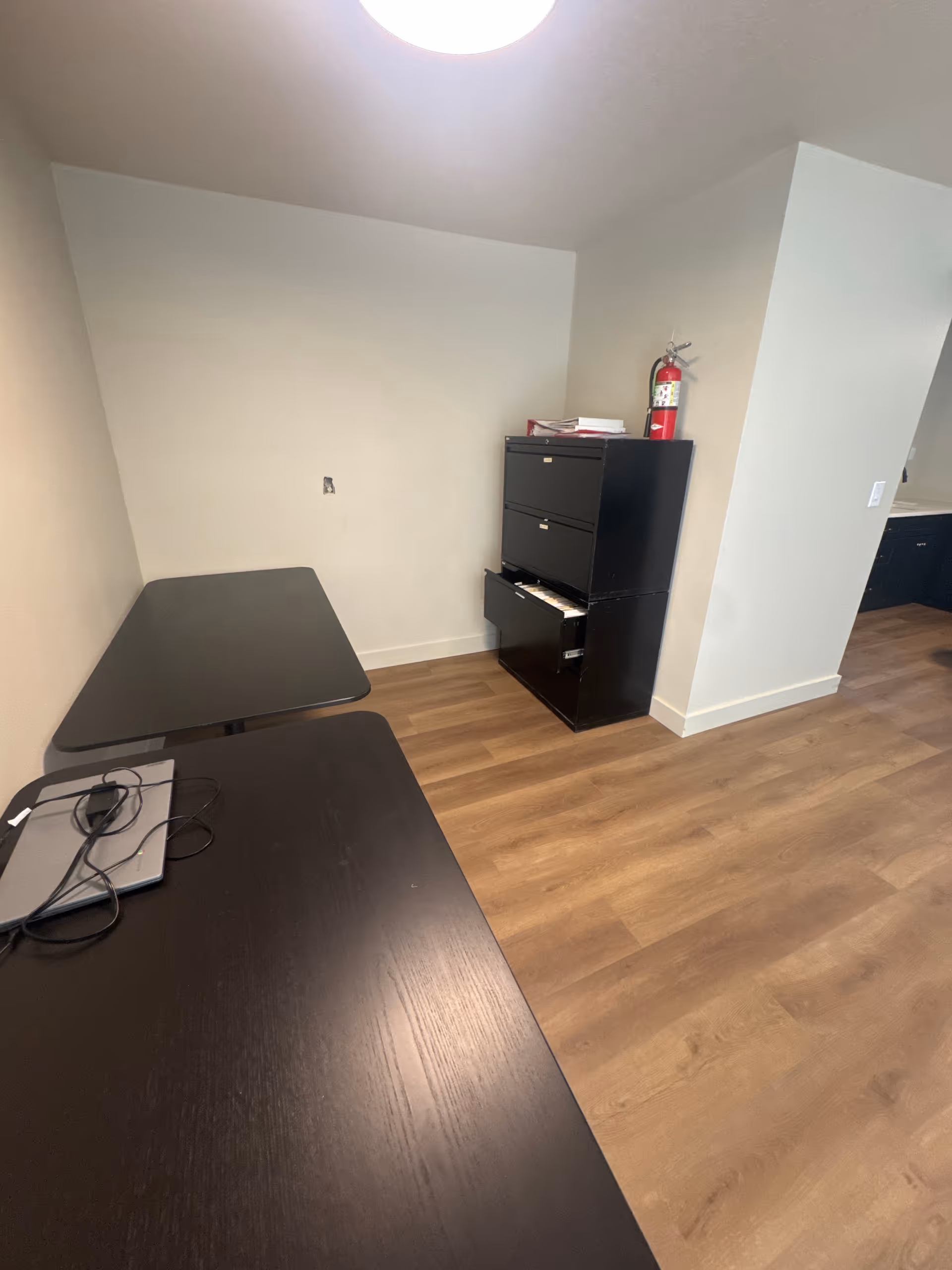 Small staff office with two dark desks, a filing cabinet, laptop, cords, and a fire extinguisher.