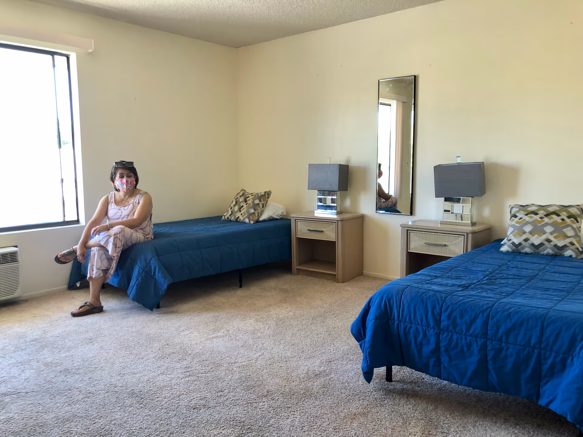 Resident wearing a pink mask sits on a bed in a two-bed senior living bedroom.