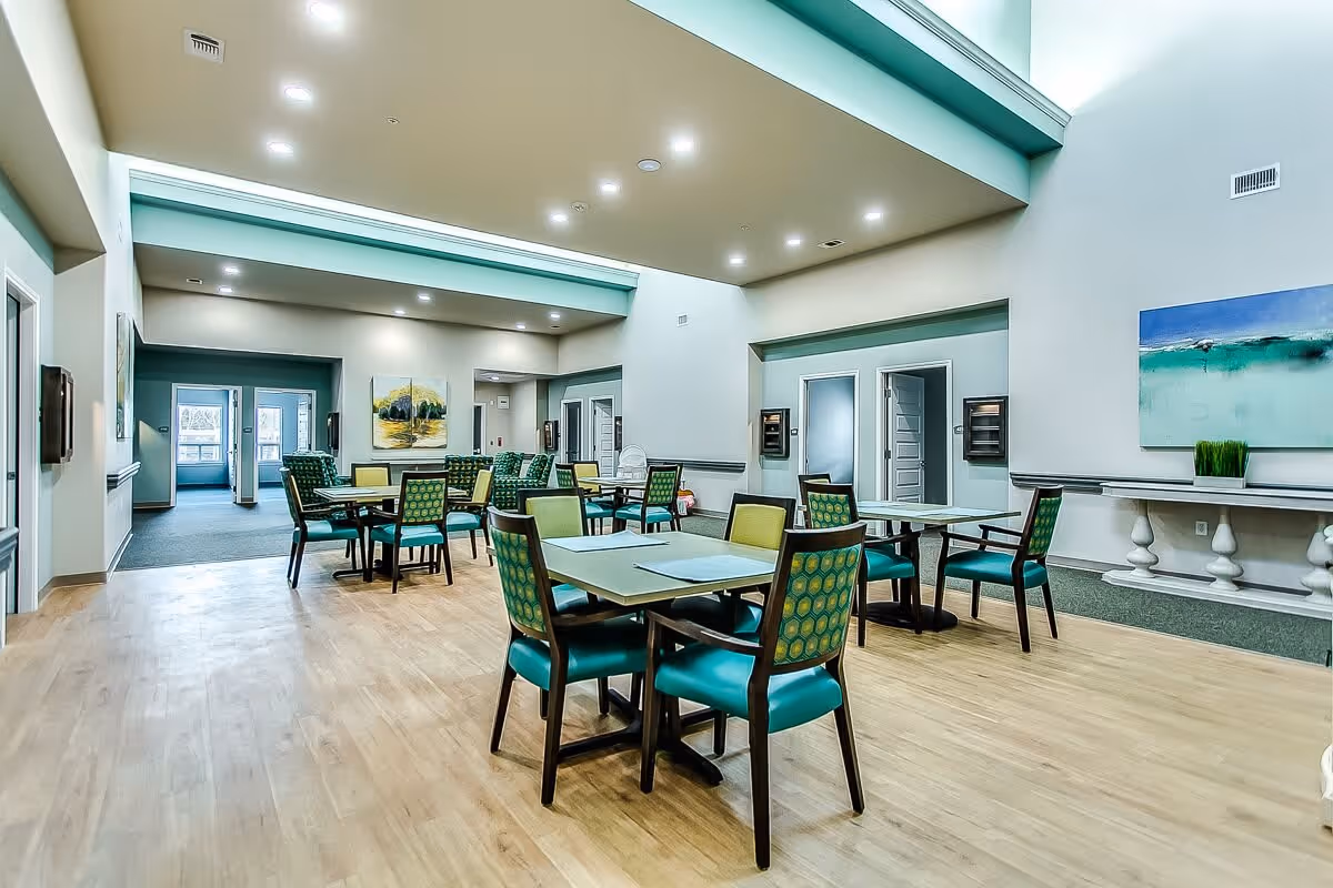 A spacious dining room in a senior living facility with multiple tables and chairs arranged neatly. The room has high ceilings with recessed lighting, light-colored walls, and wooden flooring. There are paintings on the walls and a decorative console table with a plant. Several doorways lead to other rooms.