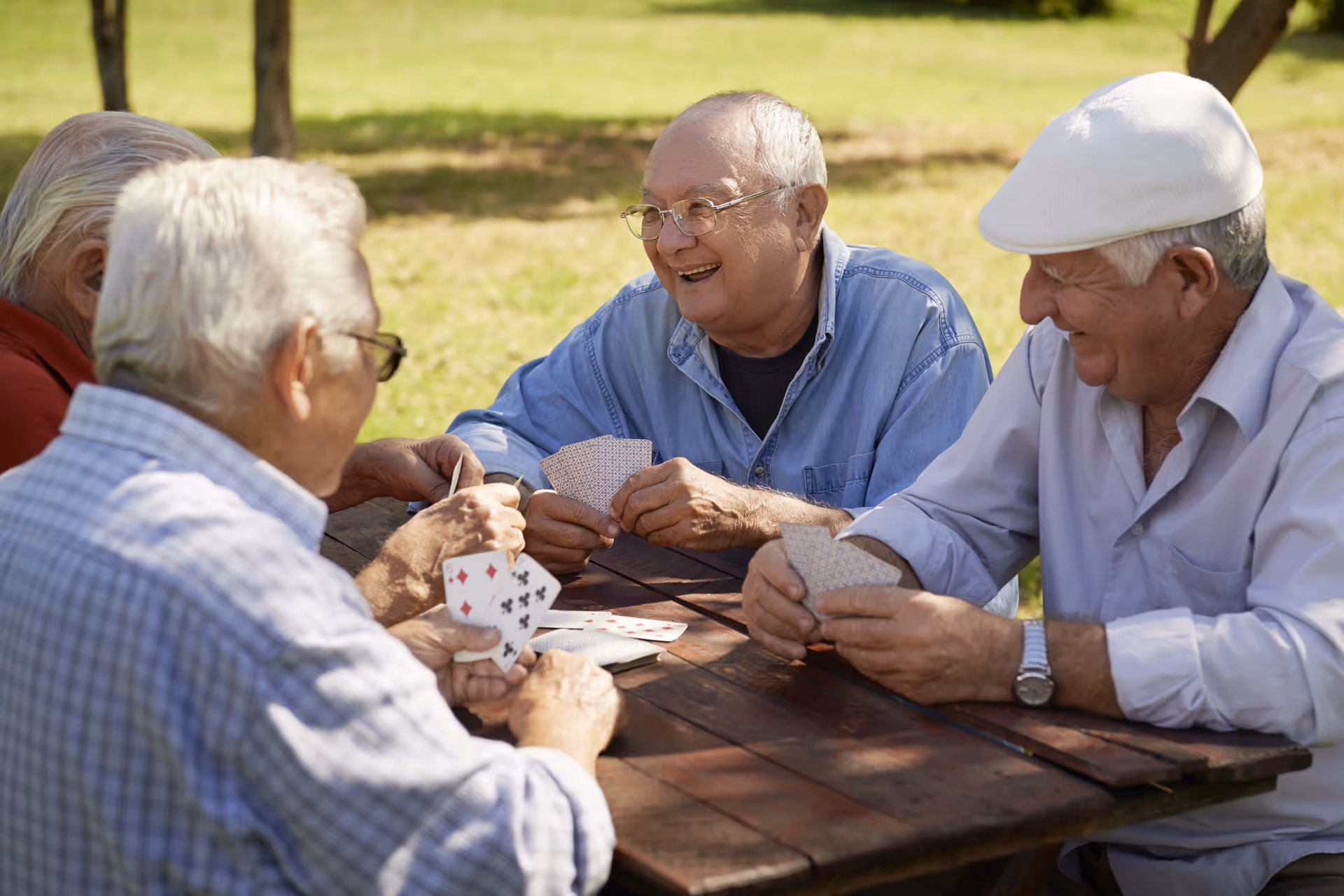 Senior men enjoy a card game at an outdoor wooden table in a sunny park.