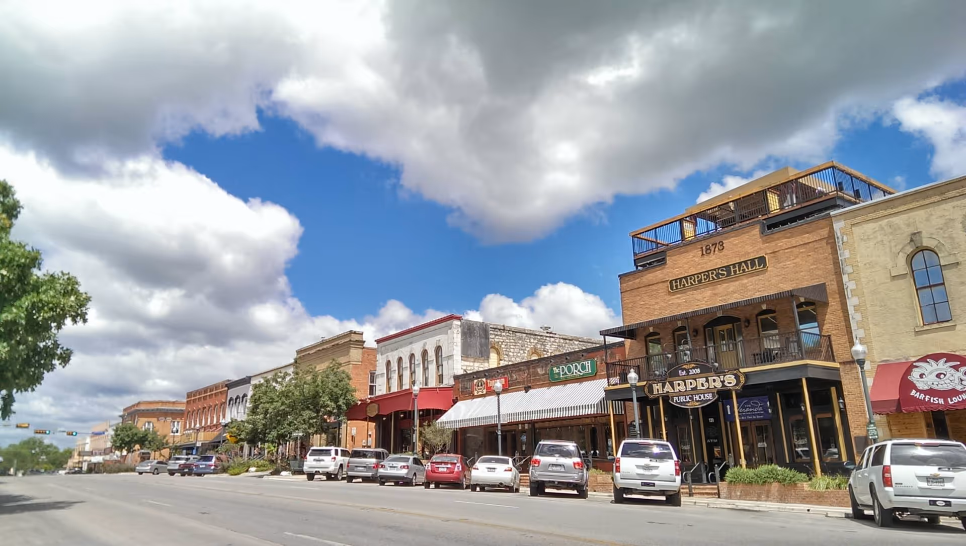 A small-town street with brick storefronts, cars parked along the road, under a blue sky.
