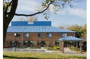 Brick facility with blue metal roof and entry canopy, surrounded by trees and a parking lot.