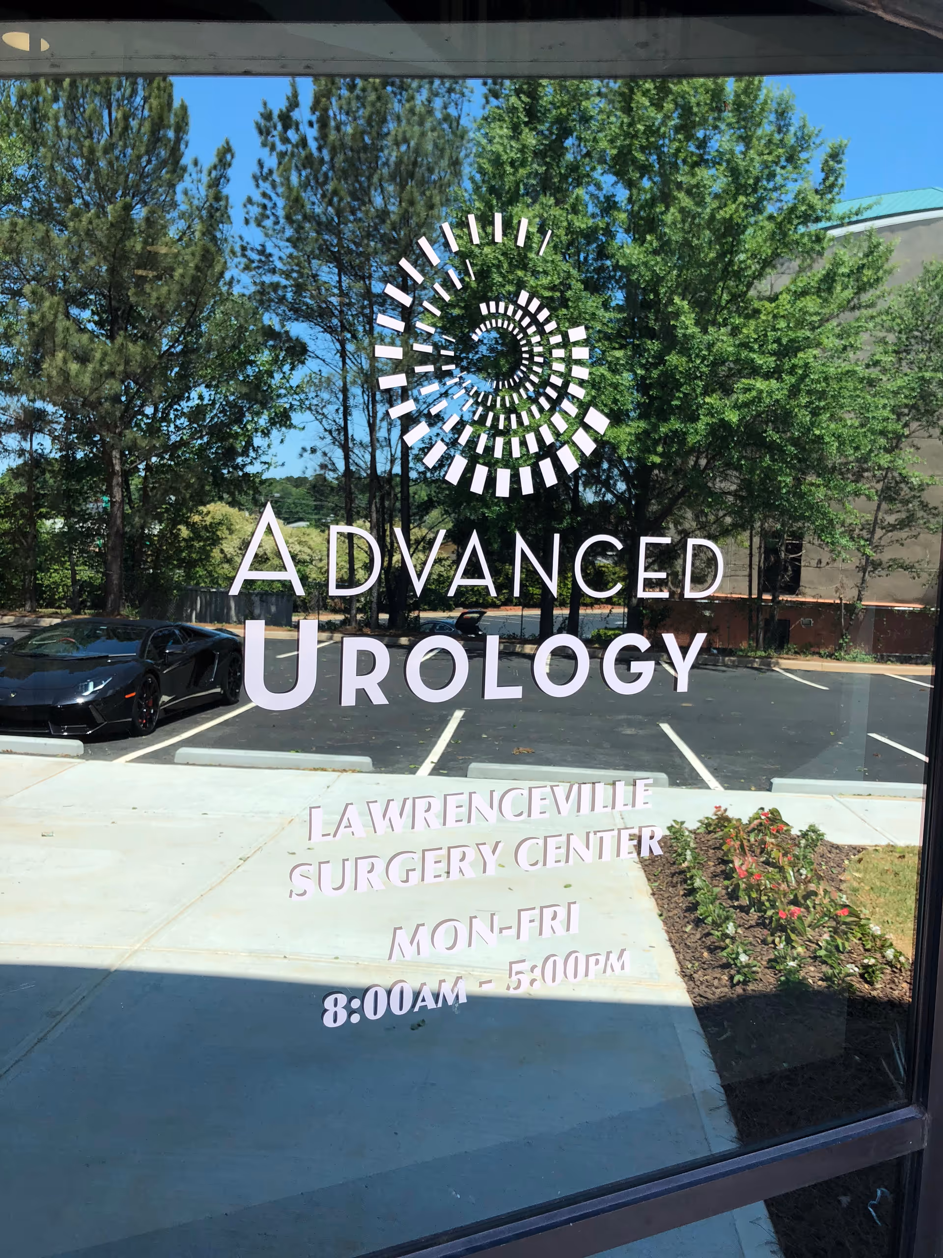 Glass storefront door shows Advanced Urology signage with Lawrenceville Surgery Center, parking lot and greenery.