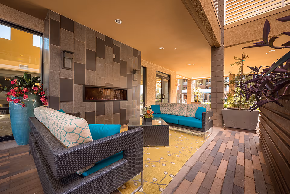 Covered outdoor seating area with wicker sofas, turquoise cushions, a wall-mounted fireplace, and potted plants.