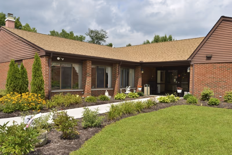 Exterior view of a single-story brick healthcare facility with a tan shingled roof, large windows, and a covered entrance. The building is surrounded by a well-maintained garden with green shrubs, yellow flowers, and a neatly trimmed lawn under a partly cloudy sky.
