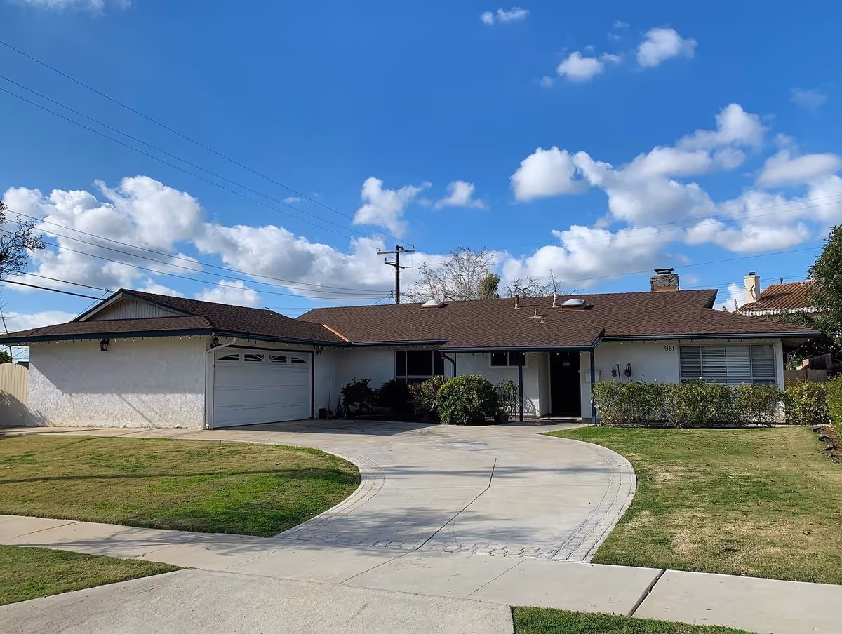 Single-story residential building with a brown shingle roof, white exterior walls, a two-car garage, and a curved concrete driveway. The front yard has green grass and bushes, and the sky is blue with scattered clouds.
