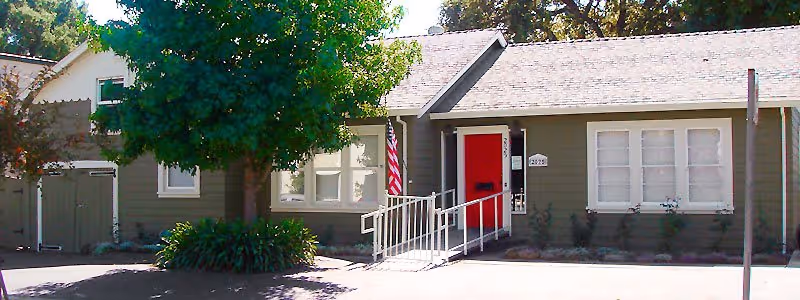 Single story senior living home exterior with red door, white railing, flag, and greenery.