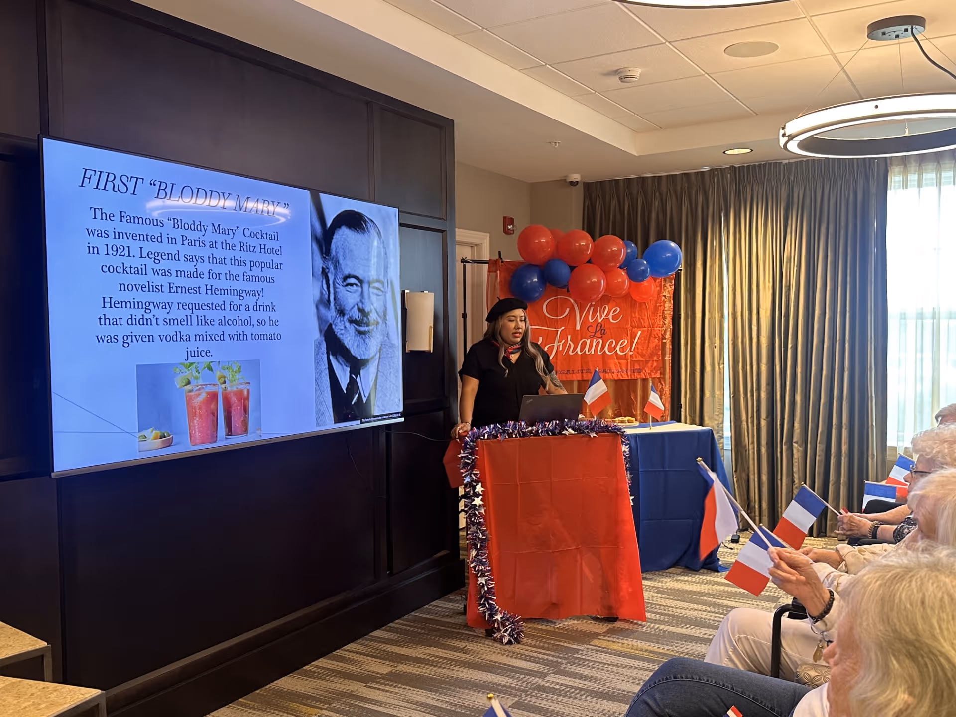 A woman stands behind a podium decorated with red cloth and tinsel, giving a presentation about the history of the Bloody Mary cocktail. The presentation slide shows a photo of Ernest Hemingway and text about the cocktail's origin. The room is decorated with red and blue balloons and a banner that reads 'Vive la France!'. Several elderly people are seated, holding small French flags and watching the presentation.