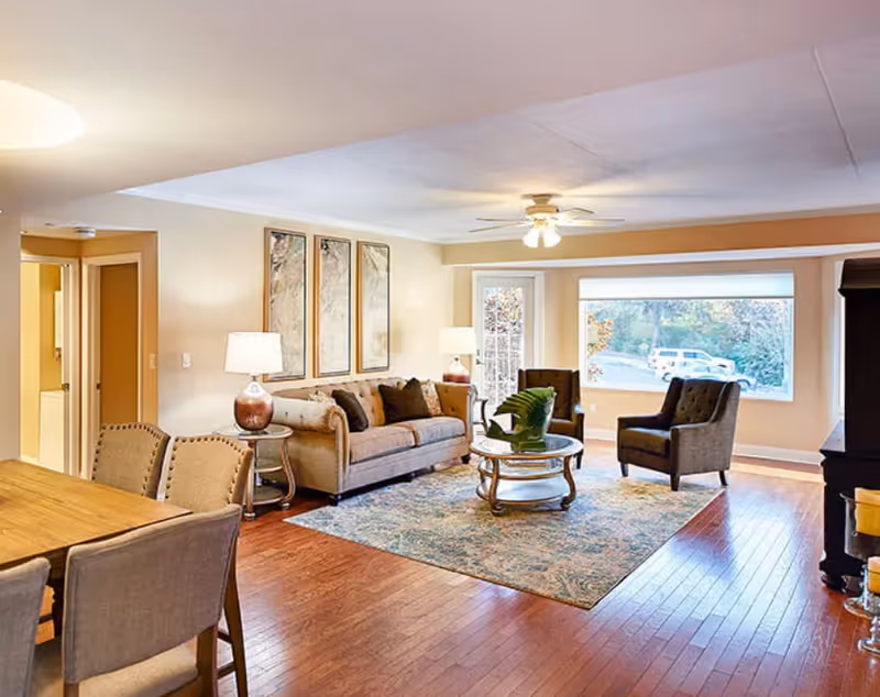 A bright and spacious living room with hardwood floors, a beige sofa with dark cushions, two armchairs, a round glass coffee table with a plant centerpiece, a patterned area rug, and large windows letting in natural light. There is a ceiling fan with lights, a dining table with chairs in the foreground, and a door leading outside.