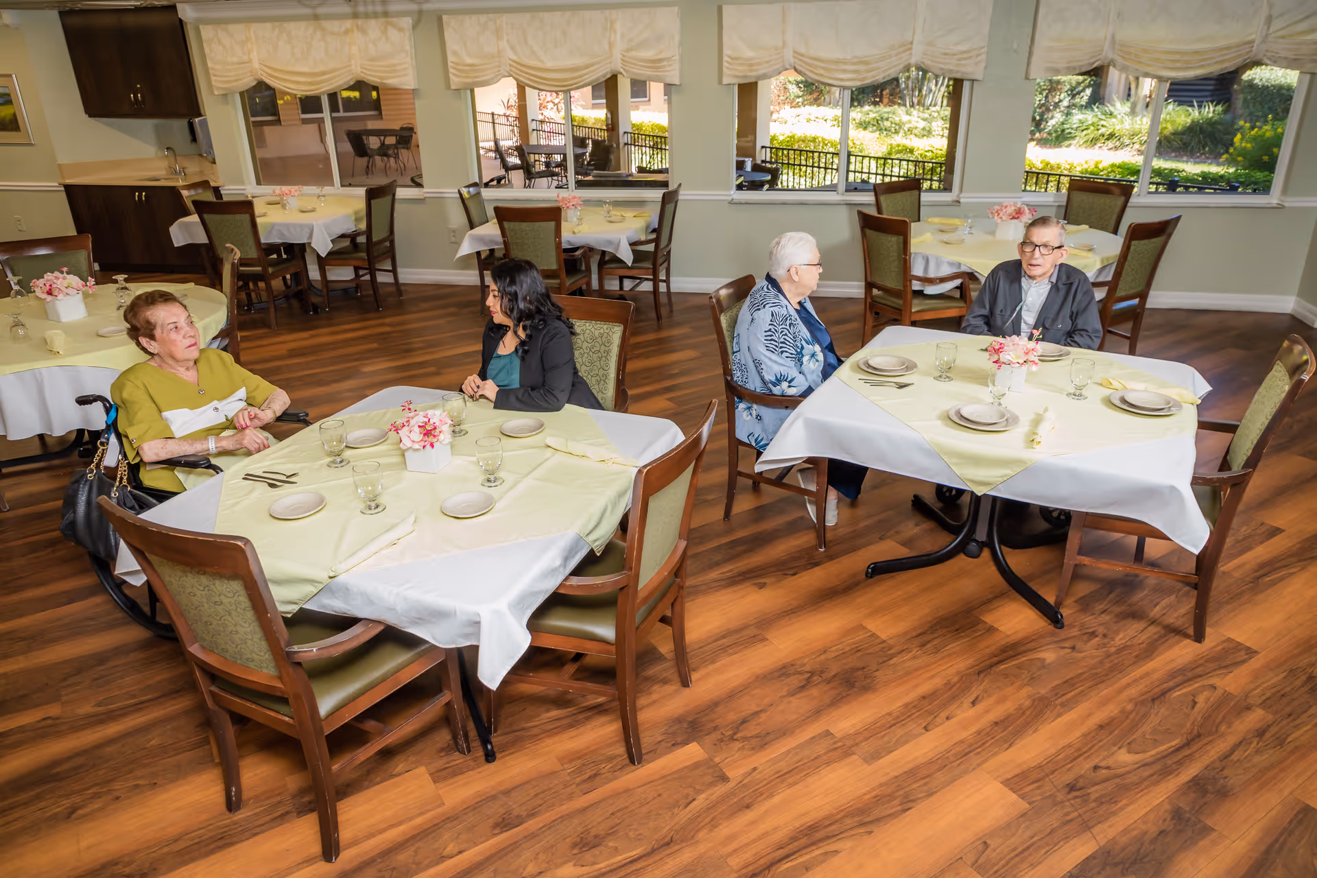 A dining room in a senior living facility with wooden floors and several tables covered with white and light green tablecloths. Four people are seated at two tables, engaged in conversation. The room has large windows with white curtains, allowing natural light to brighten the space. Each table is set with plates, glasses, and napkins, and decorated with small flower arrangements.