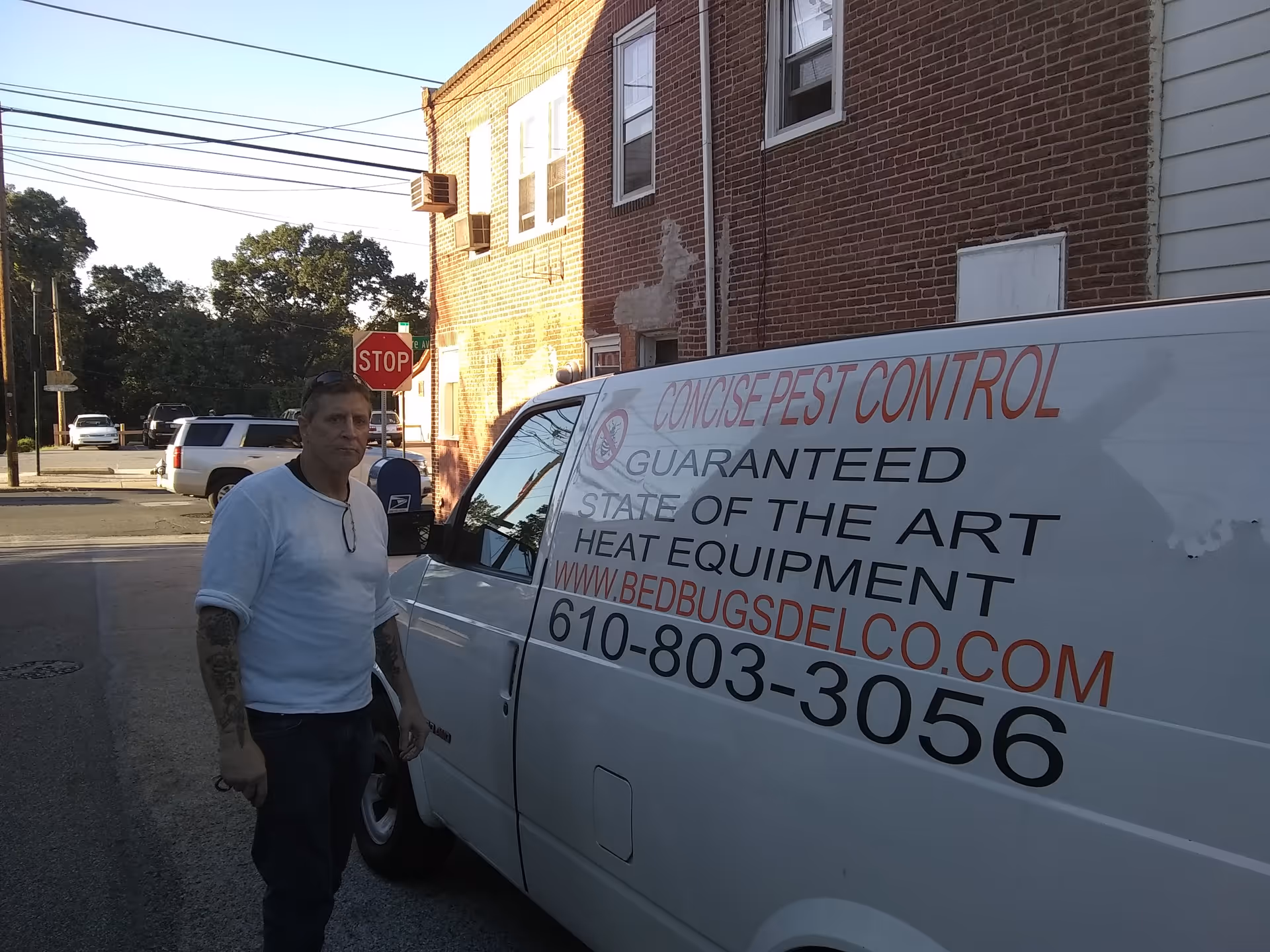 Man stands beside a white pest control van outside a brick building.