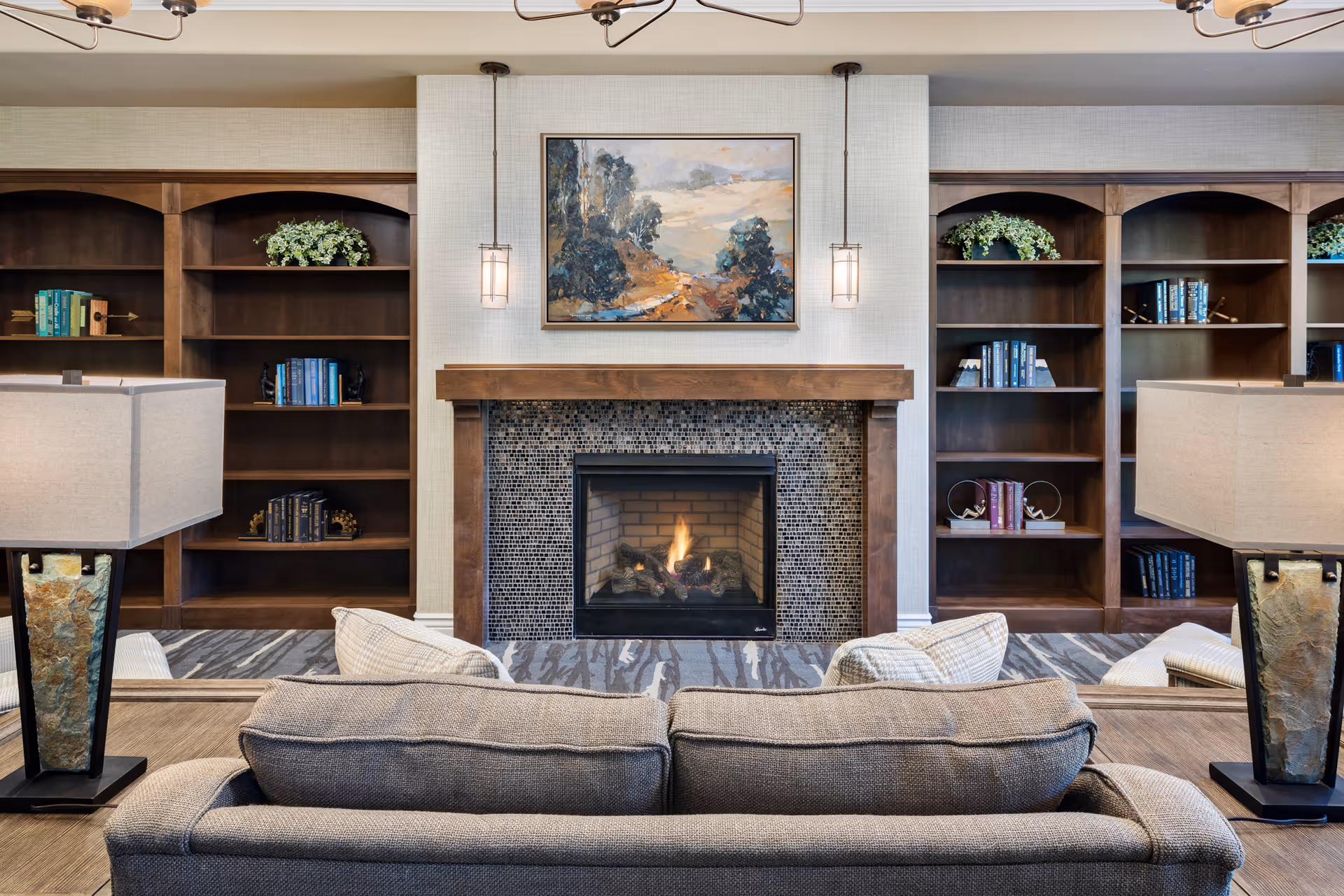 Cozy living room area with a lit fireplace in the center, flanked by built-in wooden bookshelves with books and decorative plants. A gray sofa faces the fireplace, with two table lamps on either side. Above the fireplace hangs a landscape painting, and two pendant lights hang from the ceiling.