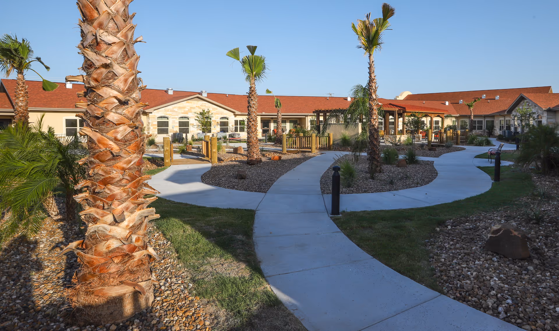 Curved concrete walkway through a landscaped courtyard with palm trees and a single-story red-roofed nursing facility in the background.