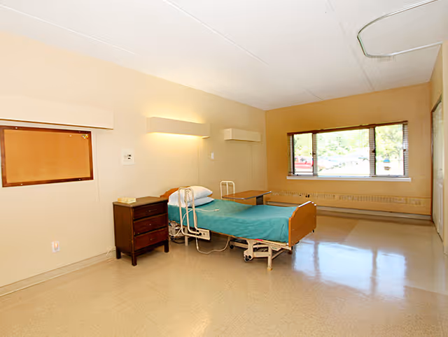 A simple, clean hospital-style bedroom with a single bed covered in a teal blanket, a wooden nightstand with a telephone, a small table on wheels, and a large window letting in natural light. The walls are beige, and the floor is a light-colored linoleum.