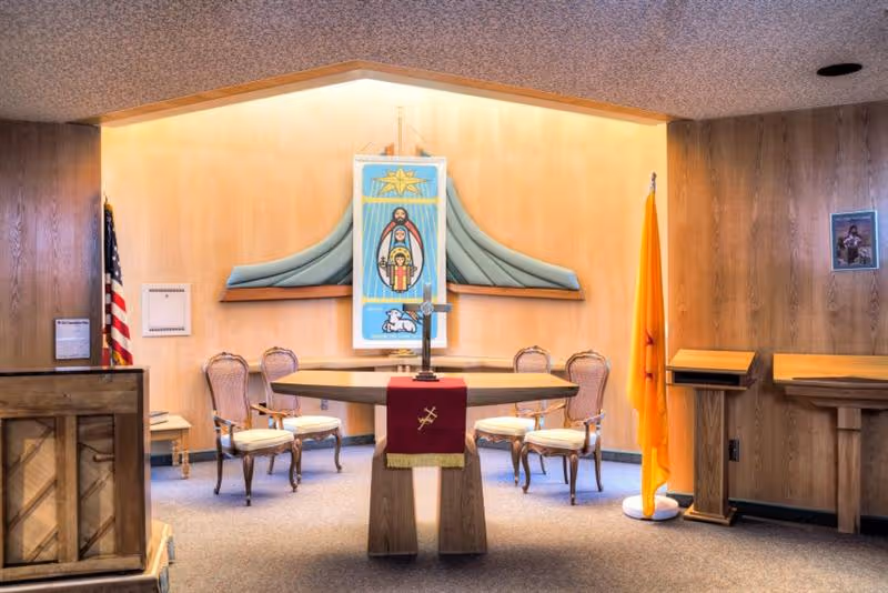 A small chapel with an altar draped in a red cloth, chairs, flags, and religious artwork on wood-paneled walls.