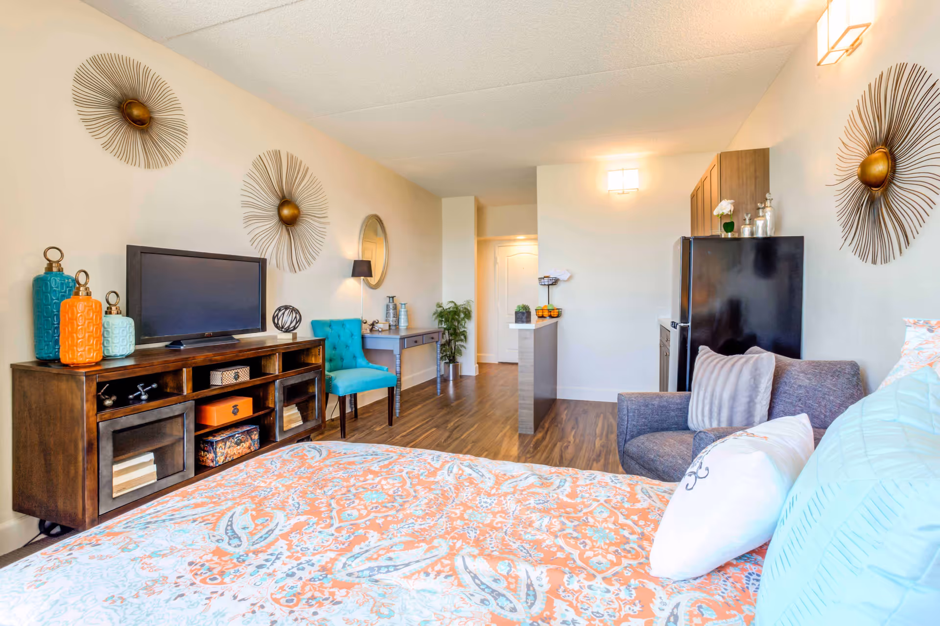 A bright and cozy senior living studio apartment featuring a bed with colorful orange and blue patterned bedding in the foreground. The room includes a gray armchair with pillows, a wooden TV stand with a flat-screen TV and decorative vases, a turquoise upholstered chair, a small desk with a lamp, and a kitchenette with a black refrigerator. The walls are decorated with three sunburst metal wall art pieces and a round mirror. The floor is wood laminate, and the room is well-lit with ceiling and wall lights.