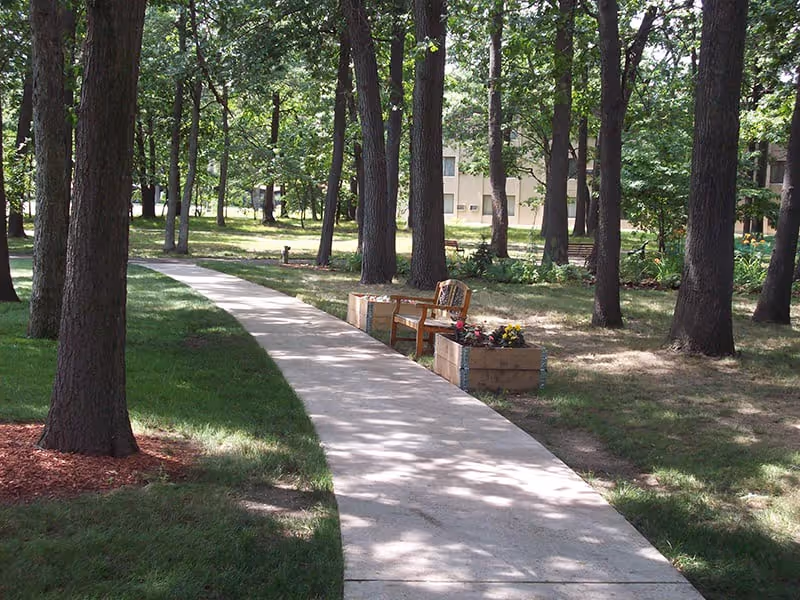 A paved walkway curves through a shaded outdoor area with tall trees and green grass. Alongside the path, there is a wooden bench with two planter boxes filled with flowers. In the background, a building is partially visible through the trees.