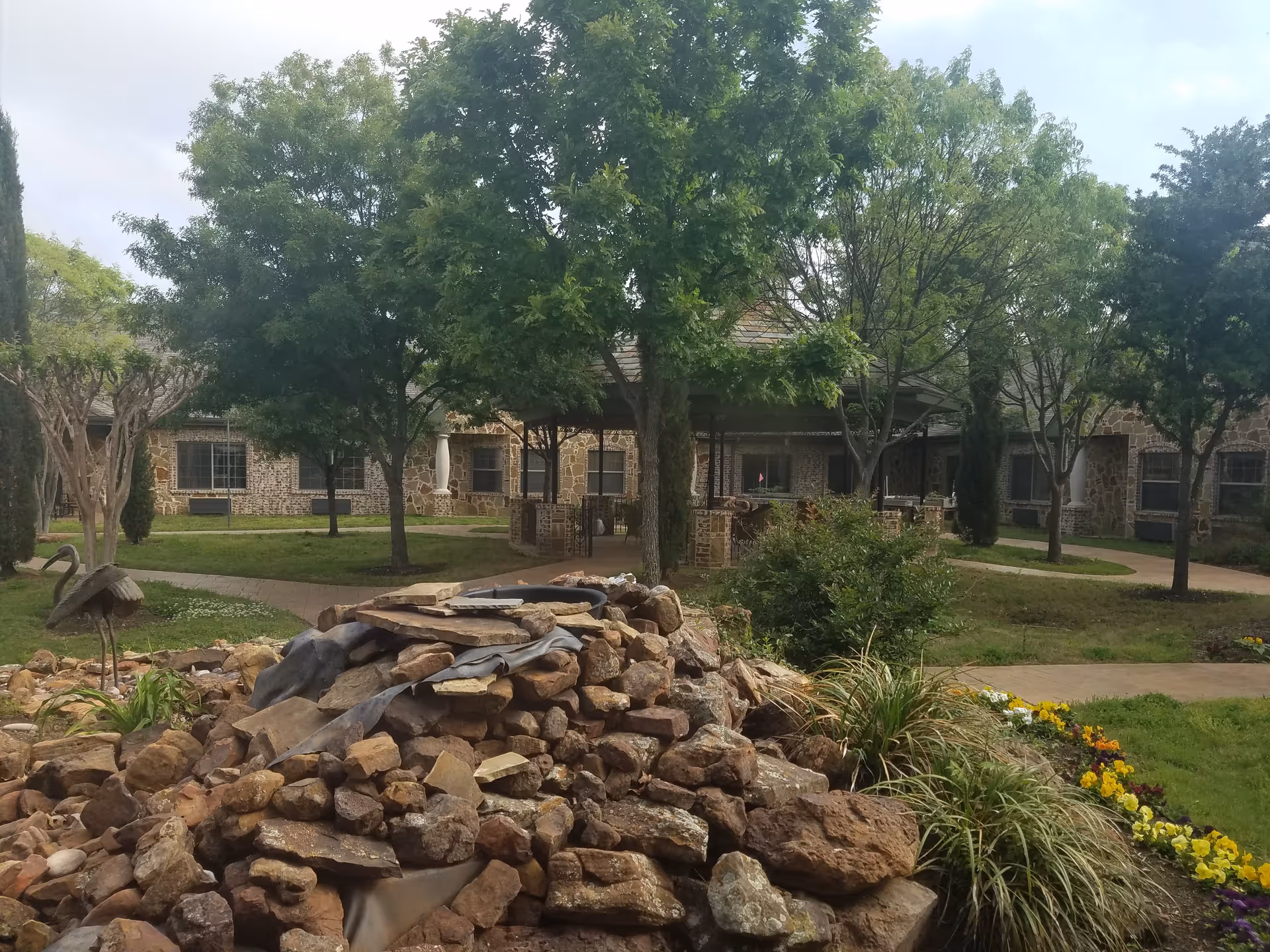Outdoor courtyard with trees, a stone fountain, and a brick building in the background.