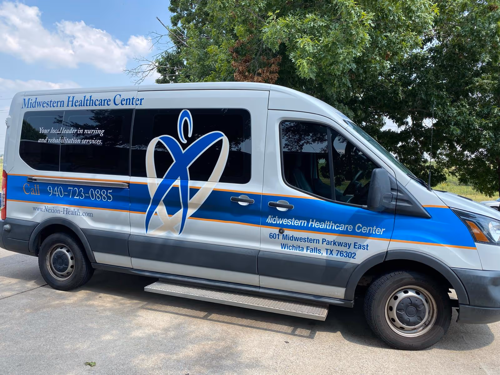 A Midwestern Healthcare Center shuttle van parked outdoors beneath trees showing the facility logo and contact information on its side.