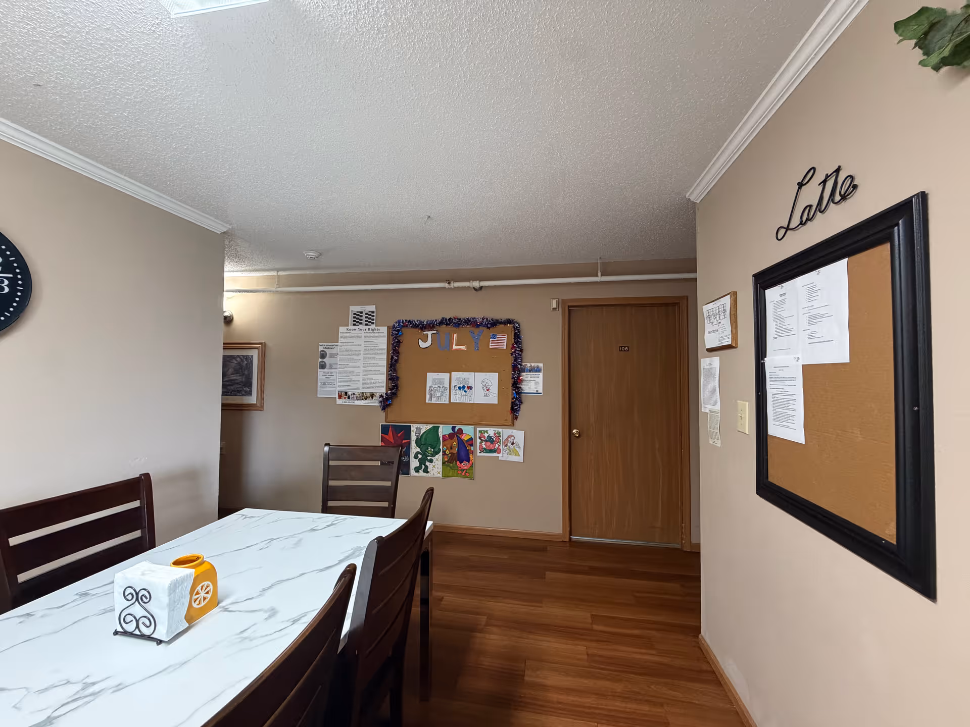 A dining room with a marble-pattern table, wooden chairs, and bulletin boards on beige walls.
