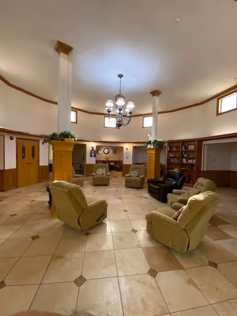 A spacious, circular common area with beige tiled flooring and cream-colored walls. The room features several comfortable armchairs arranged in a loose circle around two white columns with wooden bases. There is a chandelier hanging from the ceiling, and built-in wooden bookshelves along the walls. Small windows near the ceiling allow natural light to enter the room.