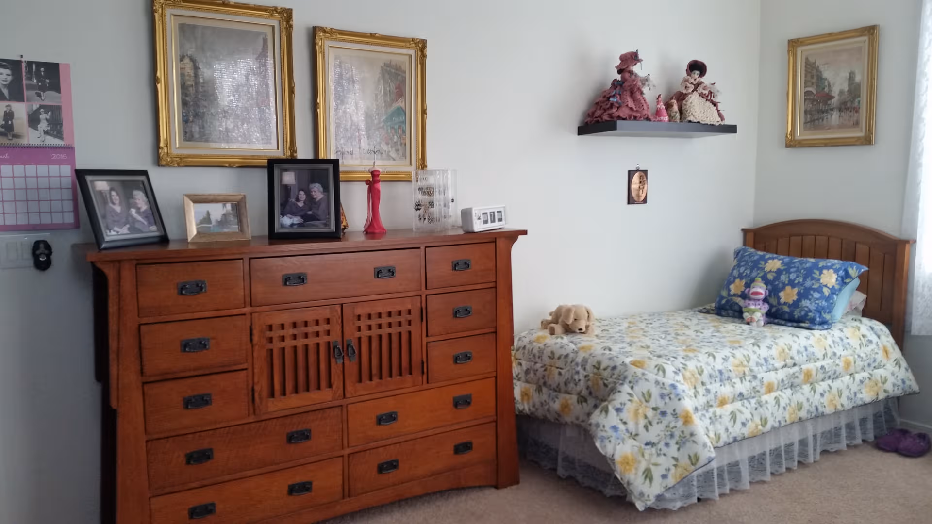 Sunlit bedroom with a wooden dresser, bed, floral bedding, and framed photos.