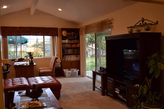 Cozy living room with a large window and sliding glass door showing a green outdoor area. The room features a wooden entertainment center with a TV, a small side table, a bookshelf filled with books and games, a desk with a chair near the window, and a brown leather ottoman in the foreground. The room is carpeted and warmly lit.