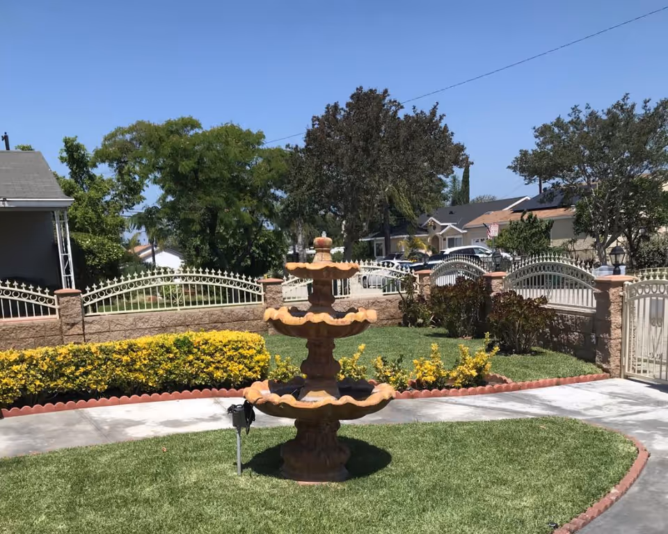 Three-tier stone fountain sits on a manicured lawn, with a gated entrance and surrounding shrubs.