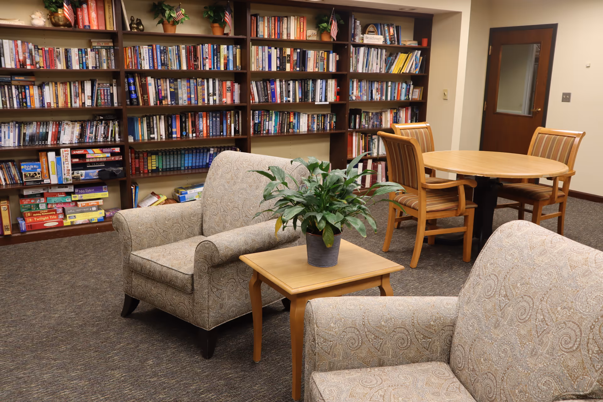 A cozy reading and sitting area in a senior living facility with two patterned armchairs around a small wooden table holding a potted plant. Behind the chairs is a large bookshelf filled with books and board games. In the background, there is a round wooden table with four striped wooden chairs and a closed wooden door with a window.