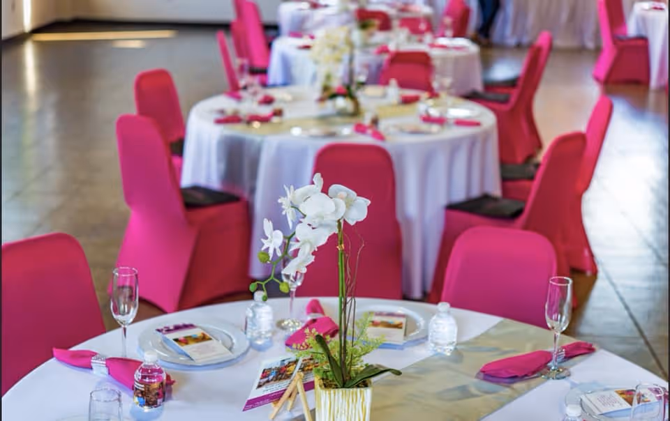 Interior banquet hall with round tables, pink chair covers, and white floral centerpieces.