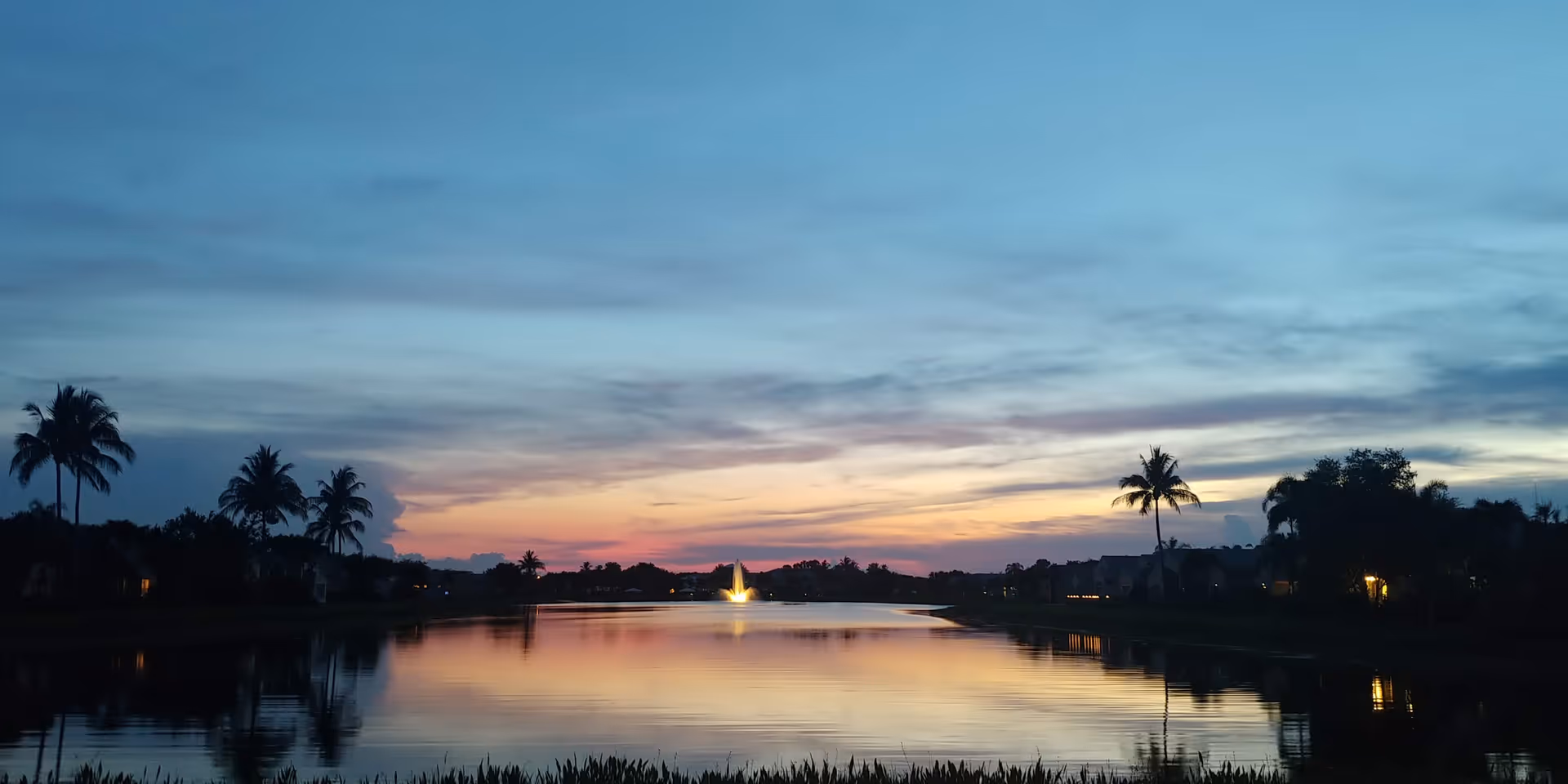 Sunset over a calm lake with palm trees and distant residential silhouettes.