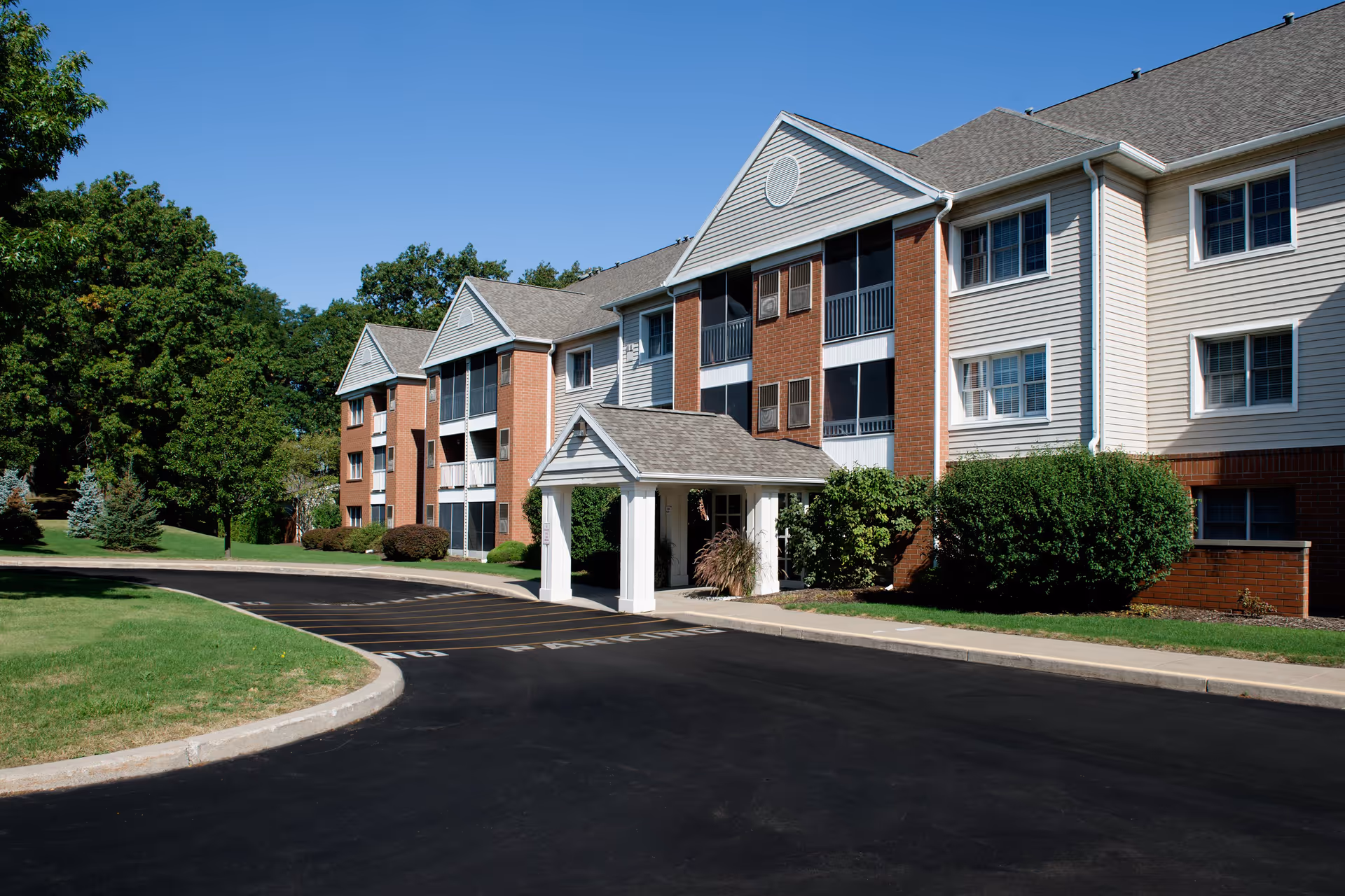 Exterior view of a multi-story senior living facility named Chapel Oaks with brick and siding facade, a covered entrance, surrounding greenery, and a clear blue sky.