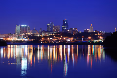 Nighttime city skyline across calm water with bright reflections.